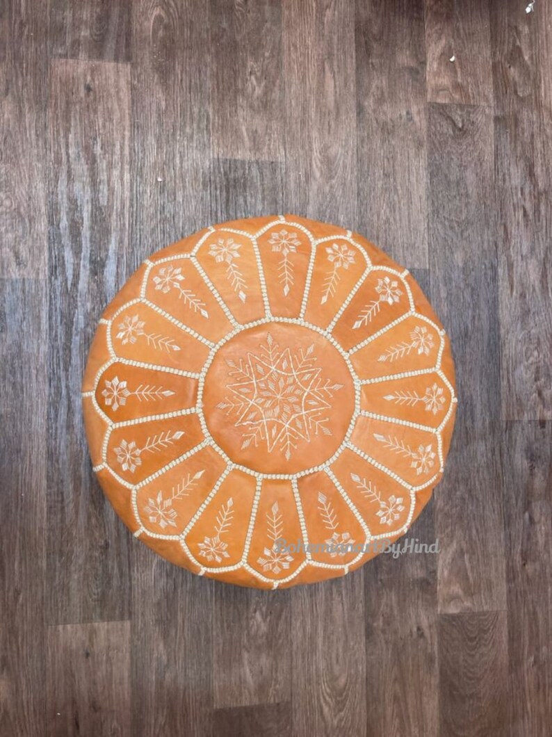 Puede incluir: Un puf redondo de cuero marr&oacute;n con dise&ntilde;os florales bordados en blanco. El puf est&aacute; sobre un suelo de madera.