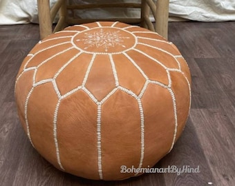 May include: A tan leather pouf with white stitching and a white snowflake design in the centre. The pouf is round and has a tufted top.