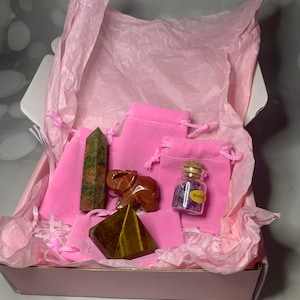 May include: A pink gift box filled with pink tissue paper and contains a green and red crystal, a brown elephant figurine, a gold pyramid crystal, and a small glass bottle with purple crystals labeled "Amethyst".