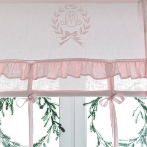 May include: Pink ruffled valance with a monogrammed wreath in the centre. The valance is made of a sheer fabric and has a delicate ruffle around the bottom edge.