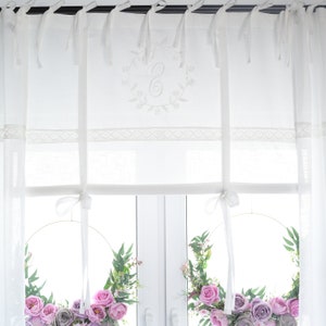 Könnte beinhalten: Weiße, durchsichtige Gardinen mit einer Spitzenborte und einem gestickten Buchstaben "E" in einem Kranz-Design. Die Gardinen sind mit weißen Bändern zurück gebunden und hinter ihnen hängen zwei Blumenkränze im Fenster.