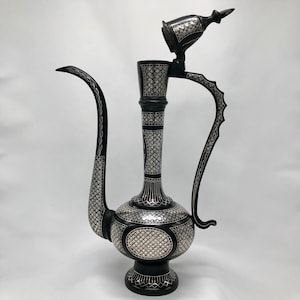 Puede incluir: Una tetera de metal adornada en negro y plata con una tapa con bisagras. La tetera tiene un cuerpo redondo y un pico largo y curvo. La tetera está decorada con intrincados patrones geométricos.