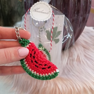 Könnte beinhalten: Ein gehäkelter Wassermelonen-Schlüsselanhänger mit grüner Schale, rotem Fruchtfleisch und schwarzen Kernen. Der Schlüsselanhänger ist an einer silbernen Kette befestigt und in einer durchsichtigen Plastiktüte mit einem weißen Etikett verpackt, auf dem "Handmade" steht.