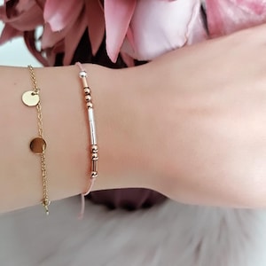 Könnte beinhalten: Zwei goldene Armbänder an einem Handgelenk. Eines hat eine feine Kette mit zwei kleinen, runden Anhängern. Das andere ist auf einem rosa Band aufgereiht und hat kleine roségoldene Perlen und einen silbernen Stab.