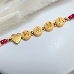 Könnte beinhalten: Ein Perlenarmband mit dem Wort "MAMA" aus cremefarbenen Buchstabenperlen. Das Armband hat einen goldenen Herzanhänger und ist mit roten, gelben und cremefarbenen Perlen aufgefädelt.