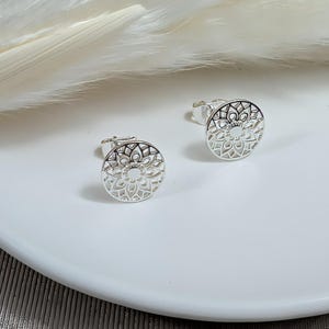 Könnte beinhalten: Ein Paar silberne Ohrstecker. Jeder Ohrstecker hat ein kreisförmiges, durchbrochenes Design mit einem floralen oder mandalaartigen Muster. Die Ohrringe werden auf einer weißen Oberfläche mit einem weichen, verschwommenen Hintergrund präsentiert.