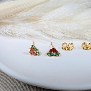 Könnte beinhalten: Goldfarbene Ohrstecker mit Wassermelonen-Scheiben-Design. Die Ohrringe haben rote und grüne Edelsteine, mit goldenen Ohrring-Verschlüssen. Die Ohrringe werden auf einer weißen Oberfläche präsentiert.