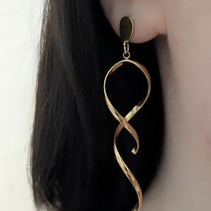 Könnte beinhalten: Goldfarbene, lange, spiralförmige Ohrhänger. Die Ohrringe haben einen kleinen, oval geformten Stecker, der mit einem geschlungenen, verdrehten Design verbunden ist. Die Ohrringe bestehen aus glänzendem, metallischem Material und hängen vom Ohr herab.