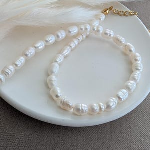Könnte beinhalten: Ein zartes Perlenketten- und Armband-Set. Die cremefarbenen, oval geformten Perlen sind aufgefädelt und erzeugen einen klassischen Look. Die Halskette hat einen goldfarbenen Verschluss und eine Kettenverlängerung. Der Schmuck wird auf einem weißen Teller präsentiert. Die Perlen messen ca. 0,5 cm.