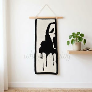May include: A rectangular crochet wall hanging featuring a black and white graphic of a ghost face mask with dripping details. The textile piece is suspended from a wooden dowel with a white cord, displaying a high-contrast pixelated design.