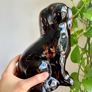 Puede incluir: Figura de perro de cerámica marrón oscuro brillante, en posición sentada. La figura tiene una superficie reflectante, con reflejos de luz y un toque de marrón. Los rasgos del perro están estilizados, y el diseño general es suave y redondeado.