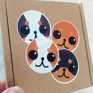 May include: Four round stickers with cartoon dog faces. Each sticker features a different colour combination of black, white, orange, and brown.