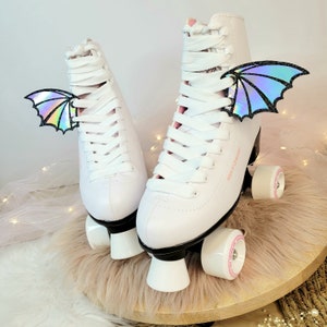 May include: White roller skates with black wheels and white platform bases. The skates have white laces and iridescent holographic wings attached to the laces.