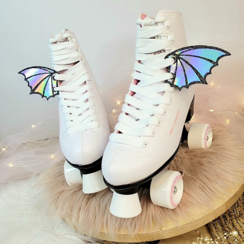 May include: White roller skates with black wheels and white platform bases. The skates have white laces and iridescent holographic wings attached to the laces.