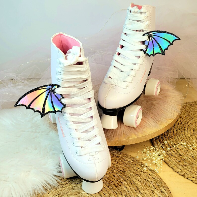 May include: A pair of white roller skates with pink accents and iridescent dragon wings attached to the laces. The skates are on a pink and brown surface.