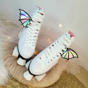 May include: A pair of white roller skates with pink accents and iridescent butterfly wings attached to the laces. The skates are on a white fluffy surface.