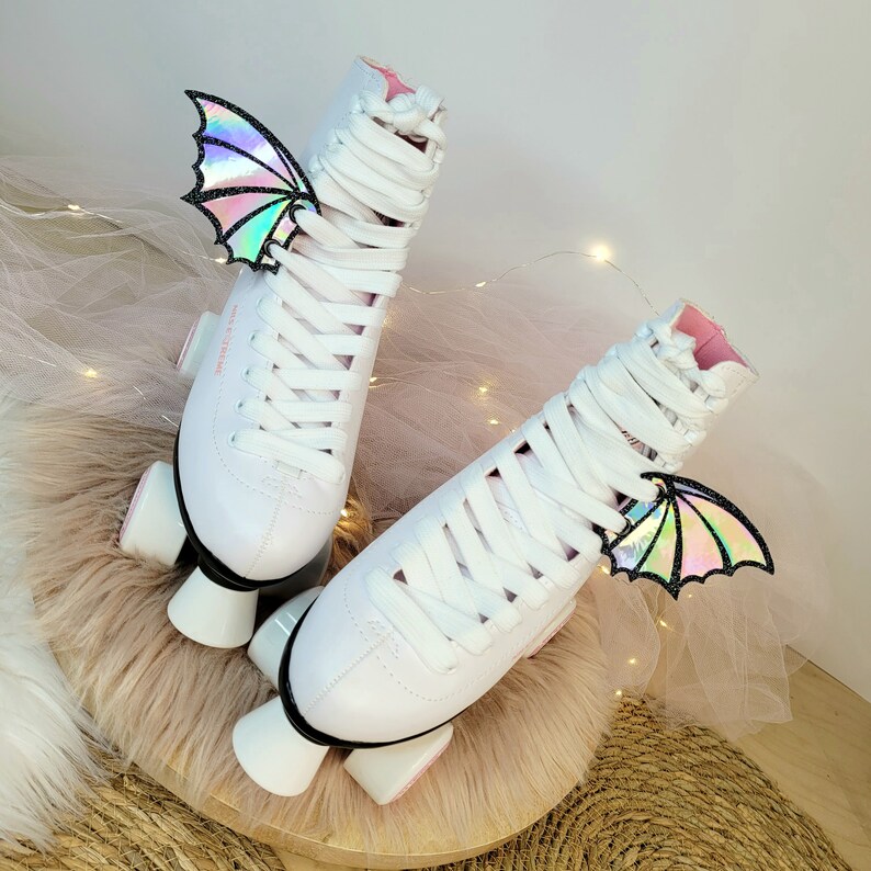 May include: A pair of white roller skates with pink accents and iridescent butterfly wings attached to the laces. The skates are on a white fluffy surface.