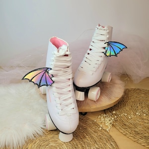 Puede incluir: Patines blancos con detalles rosas y alas iridiscentes atadas a los cordones. Los patines tienen ruedas blancas y una base blanca.