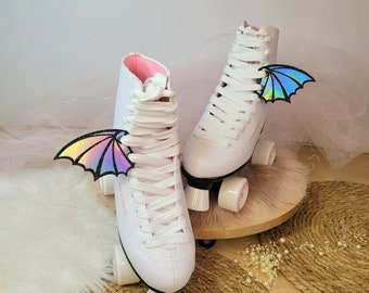 Könnte beinhalten: Weiße Rollschuhe mit rosa Akzenten und regenbogenfarbenen Flügeln, die an den Schnürsenkeln befestigt sind. Die Schuhe haben weiße Rollen und eine weiße Basis.