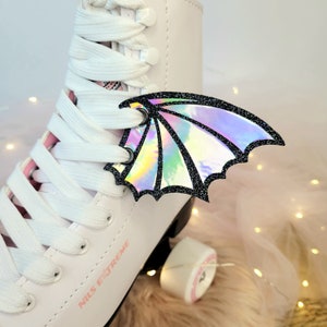 May include: White roller skates with pink laces and a holographic iridescent dragon wing charm attached to the laces. The skates have the text "NILS EXTREME" printed on the side.