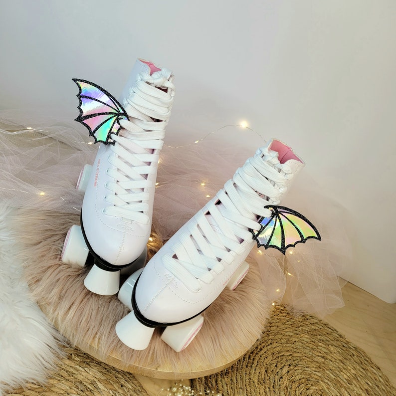 May include: White roller skates with pink accents and iridescent butterfly wings attached. The skates are on a fluffy white surface.