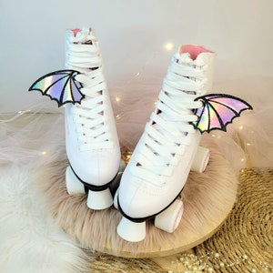 May include: A pair of white roller skates with pink laces and iridescent wings attached to the laces. The skates are on a light brown fuzzy surface.
