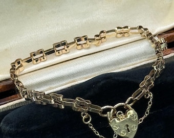 May include: A gold-toned bracelet with a heart-shaped padlock clasp. The bracelet features a delicate chain design with rectangular and decorative links. The clasp has engraved details and is connected by small chains. The bracelet is displayed in a vintage jewelry box.