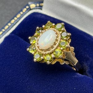 May include: A gold ring featuring a central oval opal surrounded by a halo of peridot gemstones. The ring has a detailed, ornate design with a vintage aesthetic, set against a blue velvet background.