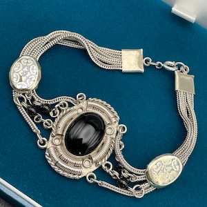 May include: A silver bracelet with a black oval gemstone surrounded by intricate silver detailing. The bracelet has multiple strands of chain and two silver charms with floral designs.