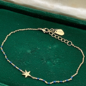 May include: A gold chain bracelet with blue beads and a gold star charm. The bracelet has a lobster clasp and a tag that says "ESTELLABARLETTI".