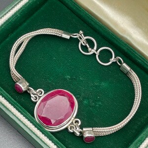 May include: A silver bracelet with a large, faceted ruby gemstone in the center. The bracelet has a braided chain design and two smaller ruby accents.