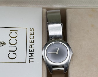 May include: A silver-toned Gucci wristwatch with a dark gray face and a light-colored leather band. The watch is displayed in a cream-colored box with a white card that reads "GUCCI TIMEPIECES".