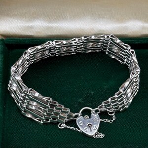 Könnte beinhalten: Ein silbernes Armband mit herzförmigem Vorhängeschloss. Das Armband besteht aus mehreren miteinander verwobenen Strängen, wodurch ein strukturiertes Aussehen entsteht. Der Verschluss ist mit einem Schlüsselloch und einer kleinen Kette versehen. Das Armband wird in einer grün mit Samt ausgekleideten Schatulle präsentiert.
