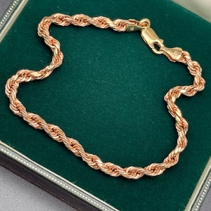 May include: A gold-colored rope chain bracelet with a lobster clasp, displayed on a dark green velvet surface within a jewelry box. The bracelet's links are tightly woven, reflecting light.