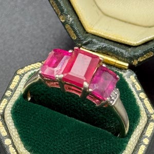 May include: A gold-coloured ring with three large, rectangular, vibrant pink gemstones. The ring is set in a classic style, with a small cluster of accent stones on one side. The ring is displayed in a vintage green jewellery box.
