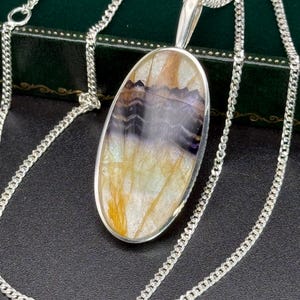 Puede incluir: Collar con colgante ovalado y cadena de plata. El colgante presenta una piedra pulida con tonos morados, amarillos y marrones. La piedra está engastada en un bisel de plata. La cadena es de estilo gourmet delicado.