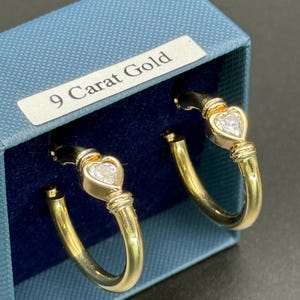 May include: A pair of gold hoop earrings with a heart-shaped clear stone. The earrings are displayed in a blue box with a label that reads "9 Carat Gold". The earrings have a polished finish and a classic design.