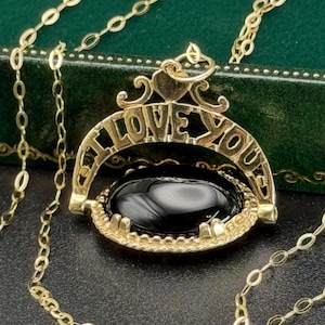 May include: A gold-coloured necklace featuring a pendant with the inscription "I LOVE YOU". The pendant has a black oval stone. The chain is a delicate link style.