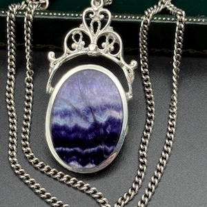 Puede incluir: Un collar con colgante de plata con una piedra ovalada reversible en tonos morados y blancos. El colgante cuelga de una cadena de plata. El colgante tiene una parte superior ornamentada y decorativa.