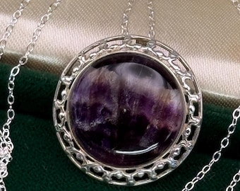 May include: A silver necklace featuring a large, round pendant with a polished, purple gemstone. The gemstone is set within an ornate, openwork silver frame. The necklace has a delicate silver chain.
