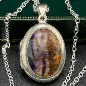 Puede incluir: Collar con colgante ovalado de plata con una piedra natural en el centro. La piedra presenta tonos de morado, marrón y dorado. El colgante cuelga de una delicada cadena de plata. El collar se muestra sobre una superficie oscura.