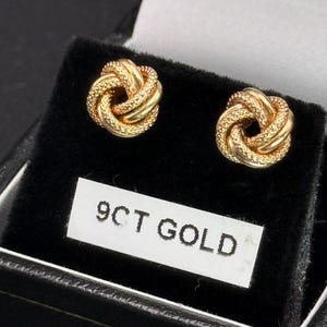 May include: A pair of gold stud earrings featuring a textured love knot design. The earrings are displayed on a black velvet insert with a white rectangular label printed with the text 9CT GOLD, indicating the material composition of the jewellery.