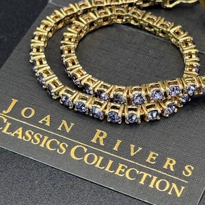 May include: A gold-tone bracelet featuring a row of purple stones. The bracelet is displayed on a black background with the text "Joan Rivers Classics Collection".