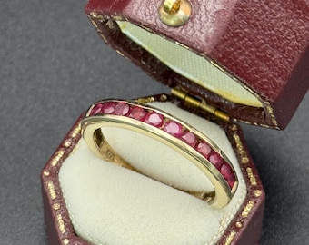 May include: A gold band ring set with a row of small, vibrant red gemstones. The ring is displayed inside a vintage burgundy leather jewelry box with a cream-colored interior.