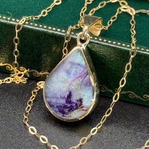 Puede incluir: Un collar dorado con un colgante en forma de lágrima. El colgante presenta una piedra brillante multicolor con tonos de morado, azul y blanco. La cadena tiene un diseño de eslabones ovalados, y el collar se muestra sobre una superficie oscura.