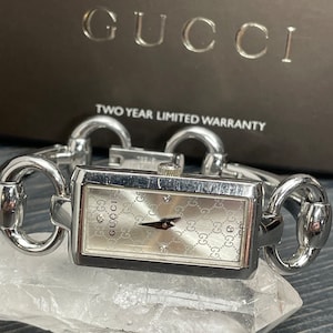 May include: A silver Gucci watch with a rectangular face and a silver chain link band. The watch face has a white background with a repeating Gucci logo pattern and diamond hour markers. The watch is on a white crystal surface. The text "TWO YEAR LIMITED WARRANTY" is visible on a brown box in the background.