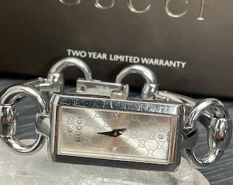 May include: A silver Gucci watch with a rectangular face and a silver chain link band. The watch face has a white background with a repeating Gucci logo pattern and diamond hour markers. The watch is on a white crystal surface. The text "TWO YEAR LIMITED WARRANTY" is visible on a brown box in the background.