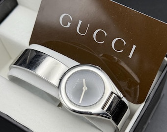 May include: A silver Gucci watch with a black face and a silver band. The watch is in a black box with a white interior. The box has a Gucci logo on the top.