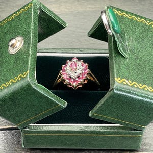 以下が含まれることがあります： ピンクの宝石が小さな白い宝石で囲まれたクラスターでセットされた、ゴールドのバンドにセットされたリング。リングは緑色のベルベットのリングボックスに展示されています。