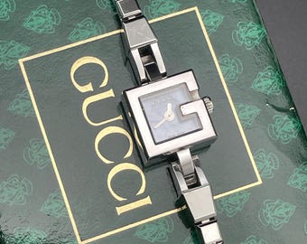 May include: A silver Gucci watch with a square face and a silver metal band. The watch is on top of a green box with the Gucci logo.